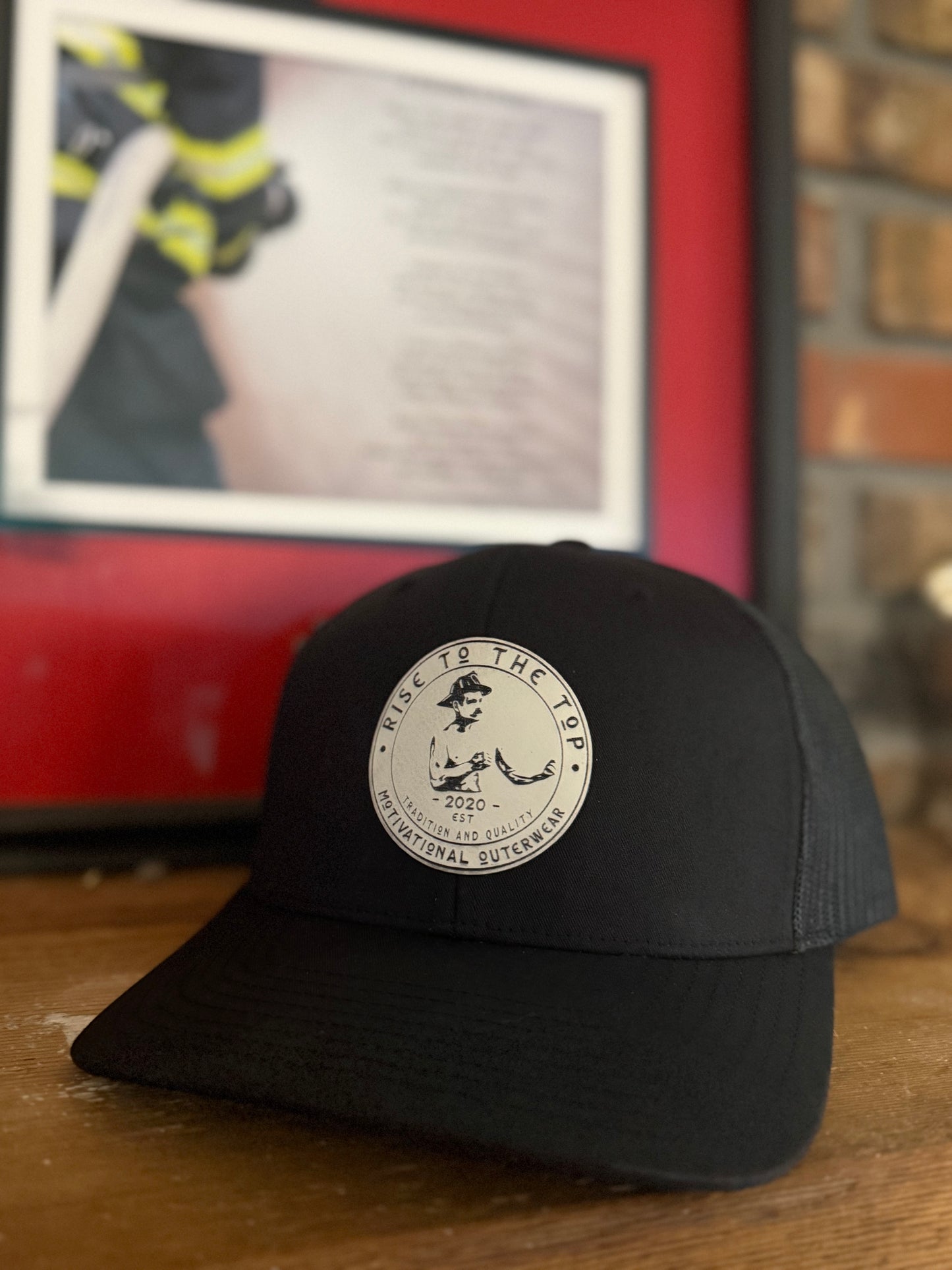 Black trucker hat with a white rise to the top patch sitting on a wood surface with a firefighter picture in the background.