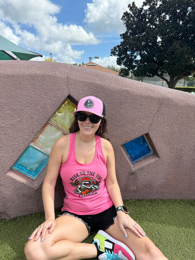 White female sitting in front of a park wall wearing a pink florida rise to the top racer tank and pink and black rise to the top trucker hat
