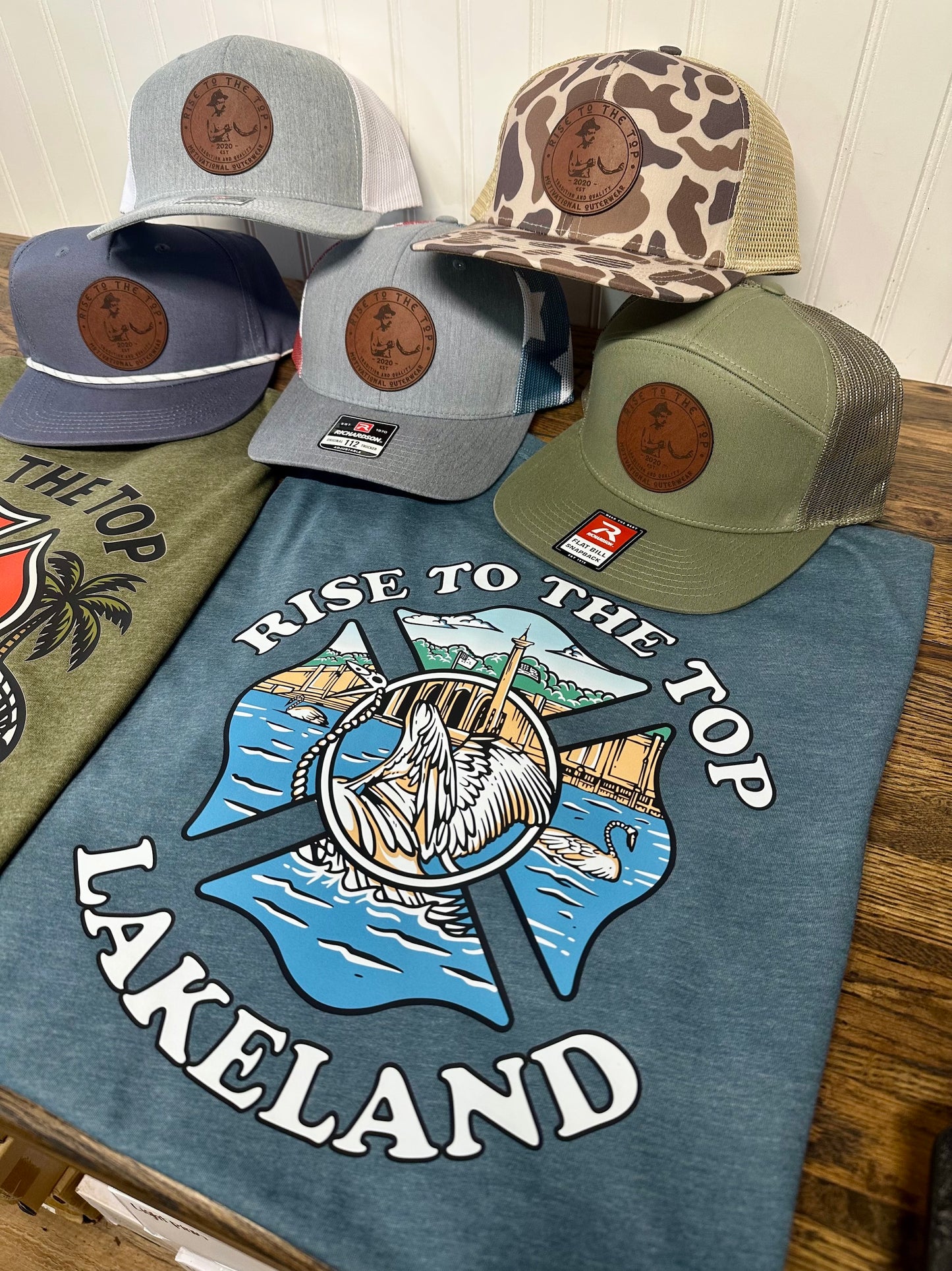 Indigo color shirt with rise to the stop lakeland swan Maltese cross logo with trucker hats in the background. 