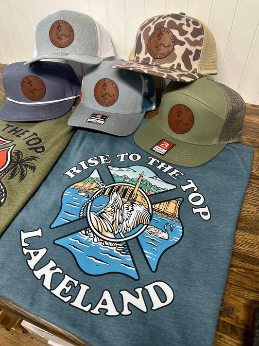 Indigo color shirt with rise to the stop lakeland swan Maltese cross logo with trucker hats in the background. 