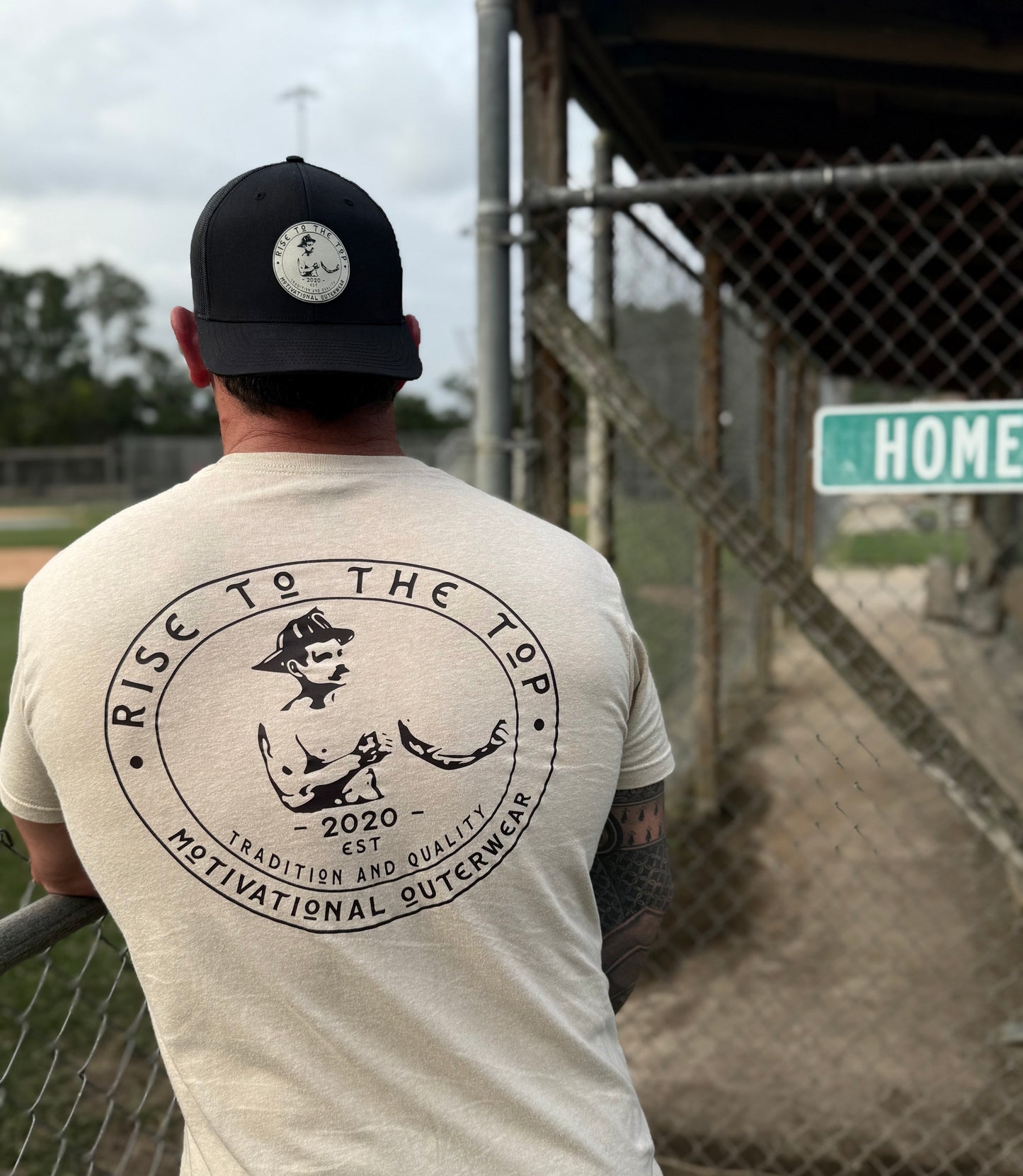 Man wearing a t-shirt with a graphic design and text (rise to the top logo), standing in front of a baseball field.