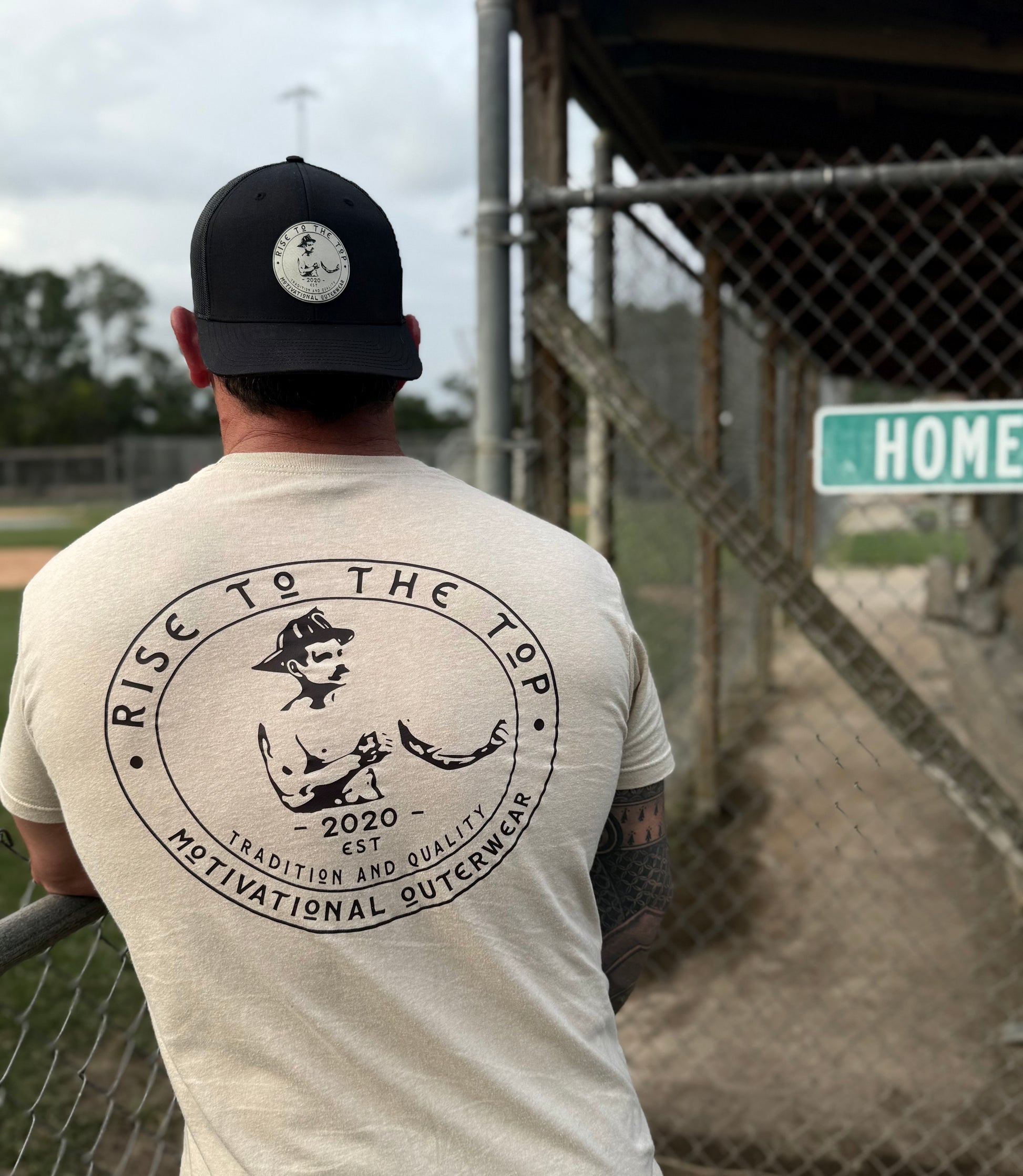 Man wearing a t-shirt with a graphic design and text (rise to the top logo), standing in front of a baseball field.